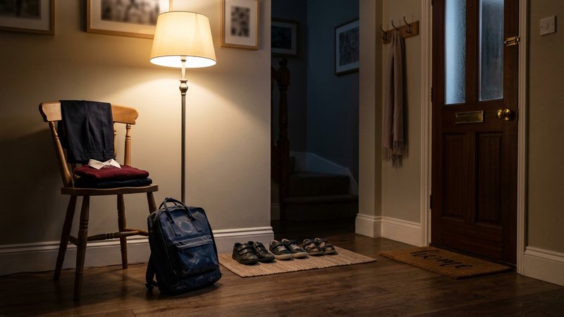 Una entrada de casa por la noche con una mochila escolar preparada junto a la puerta, el uniforme doblado sobre una sill