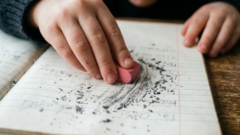Un primer plano de las manos de un niño borrando con insistencia sobre una página de cuaderno, con virutas de goma alred