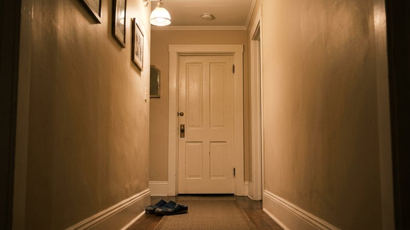 La puerta cerrada de una habitación al fondo de un pasillo de casa, con la luz cálida del pasillo iluminando el marco y